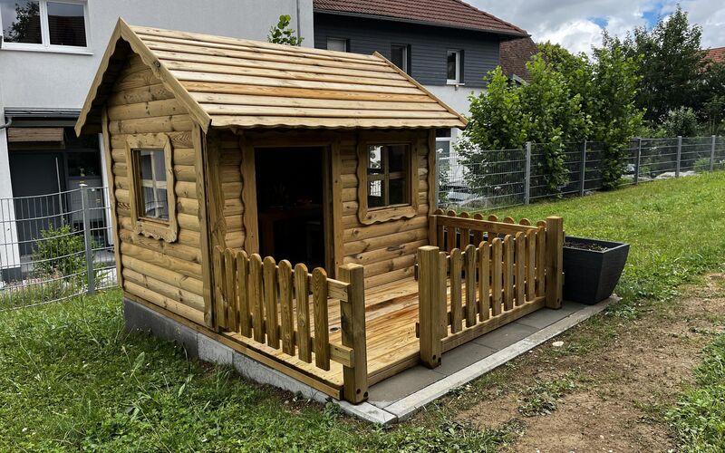 Spielhaus aus Holz mit eingezäunter Veranda auf der Wiese im Garten des Kindergartens